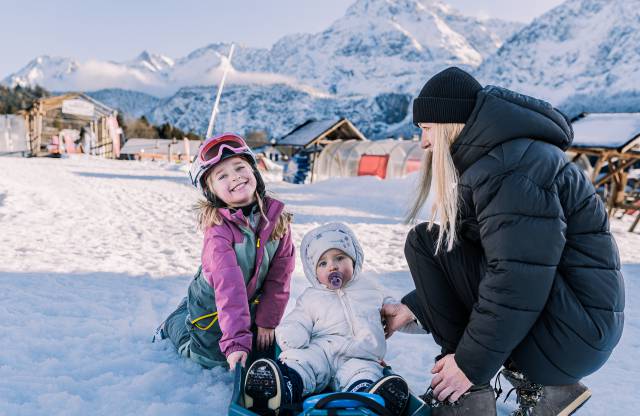 Mutter mit zwei Kindern beim Schlittenfahren im Schnee, umgeben von Bergen.