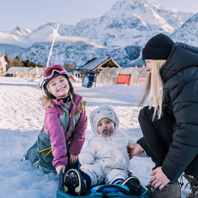 Mutter mit zwei Kindern beim Schlittenfahren im Schnee, umgeben von Bergen.