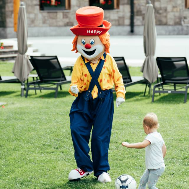 Familotel Maskottchen Happy spielt mit einem Kind Fußball im Garten des Familienhotels Tirolerhof.