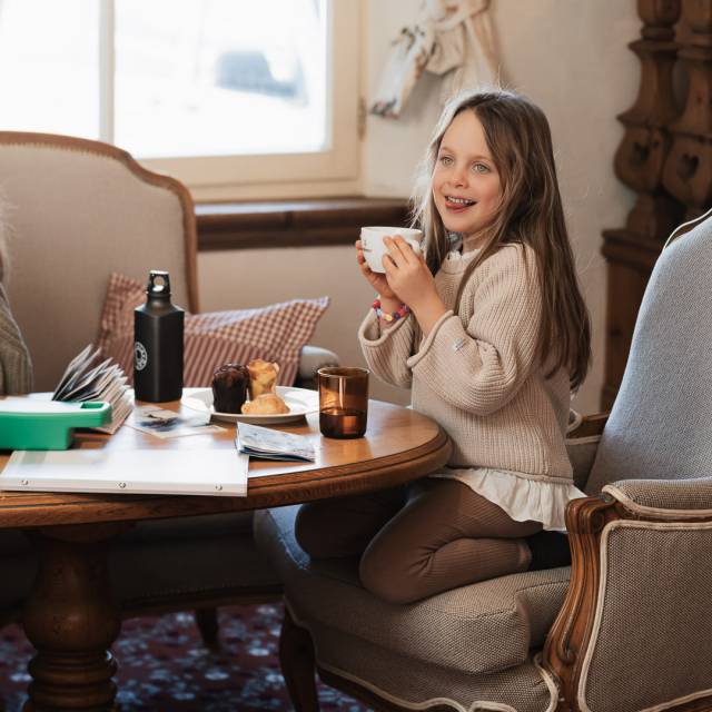 Child sitting on a cozy armchair at Tirolerhof, holding a cup in their hands.