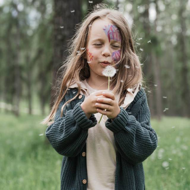 Mädchen mit Gesichtbemalung mit einer Pusteblume auf einer Wiese am Waldrand.