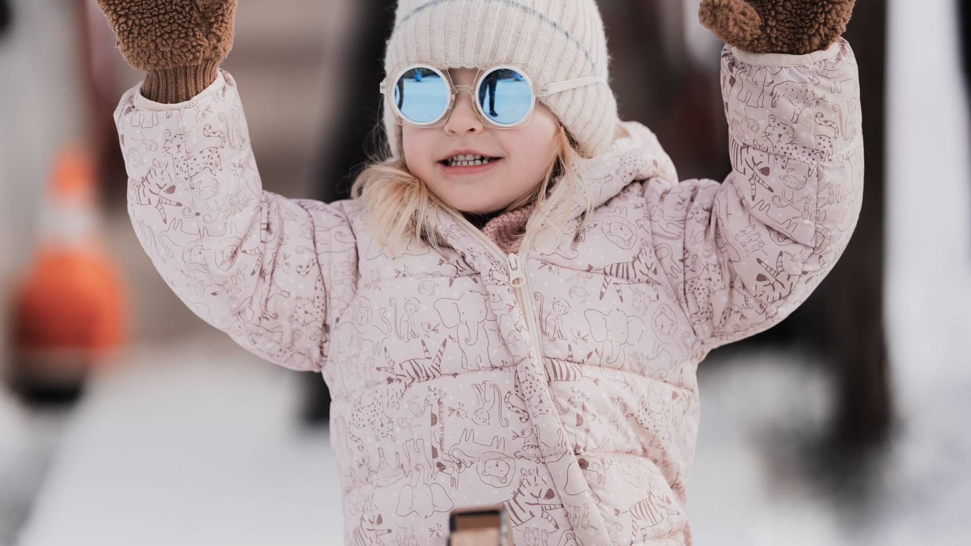 Mädchen in pinkem Skianzug und Sonnenbrille auf einem Rodel im Schnee.
