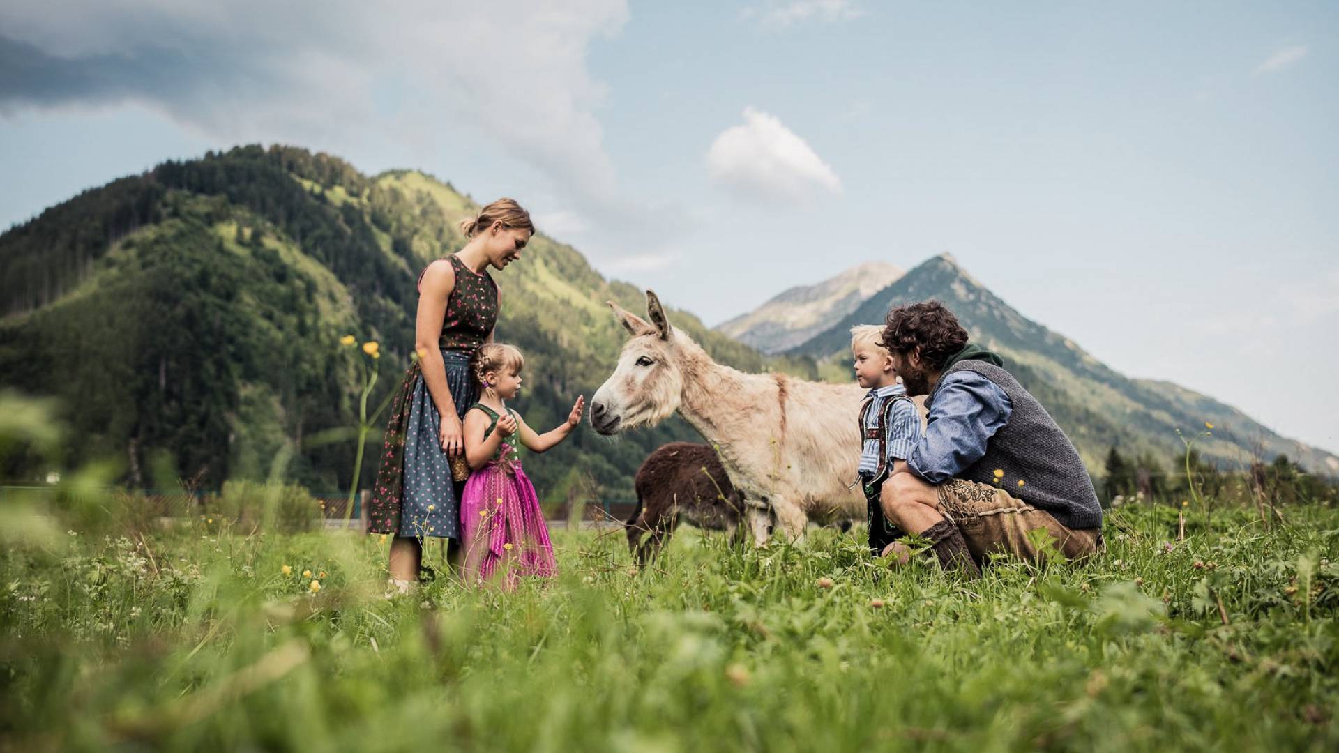 Eine Familie in traditioneller Tracht steht auf einer Wiese und streichelt einen Esel.
