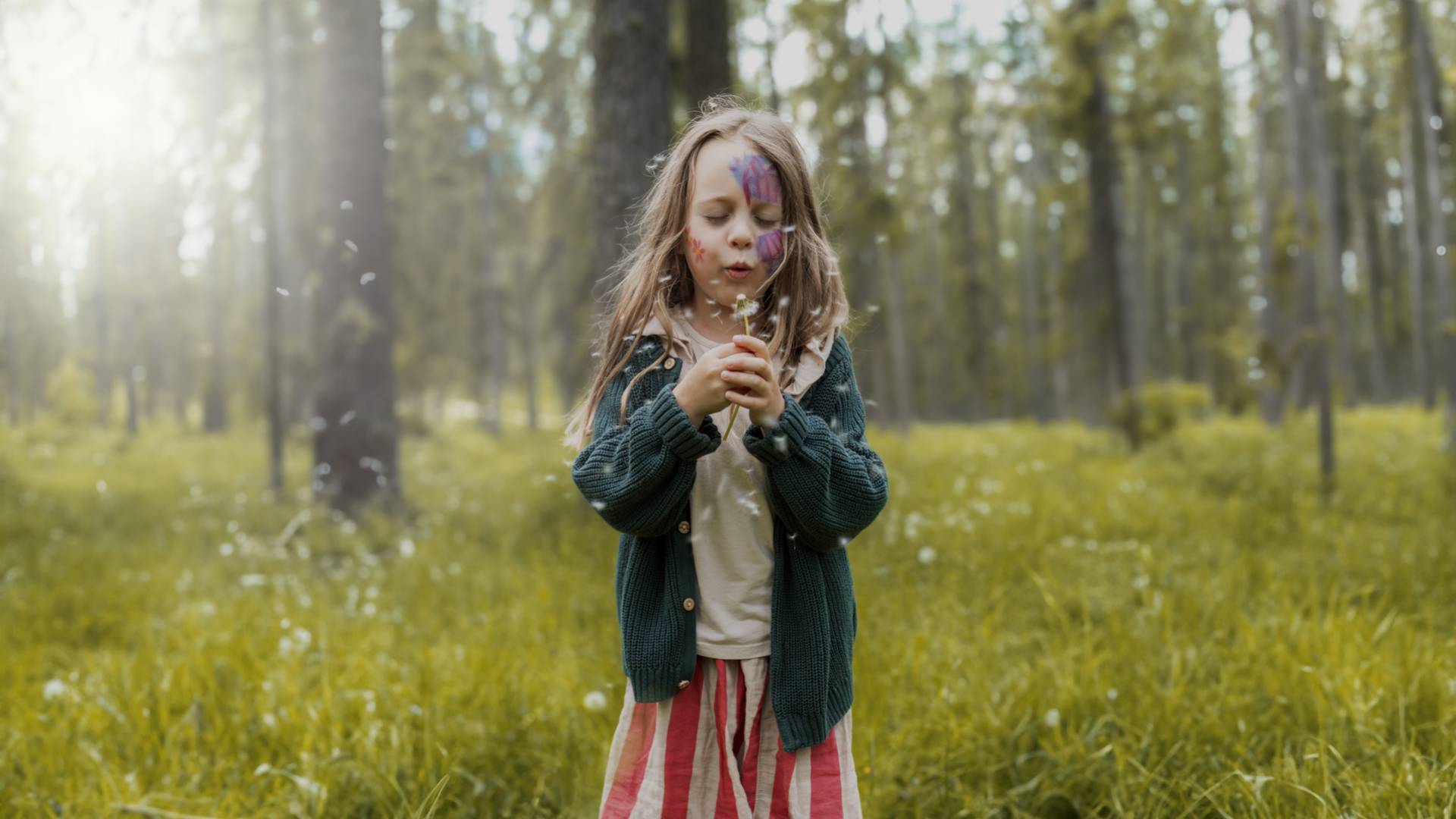 Ein natürlich gekleidetes Mädchen steht im Wald und verpustet eine Pusteblume im lichten Wald.